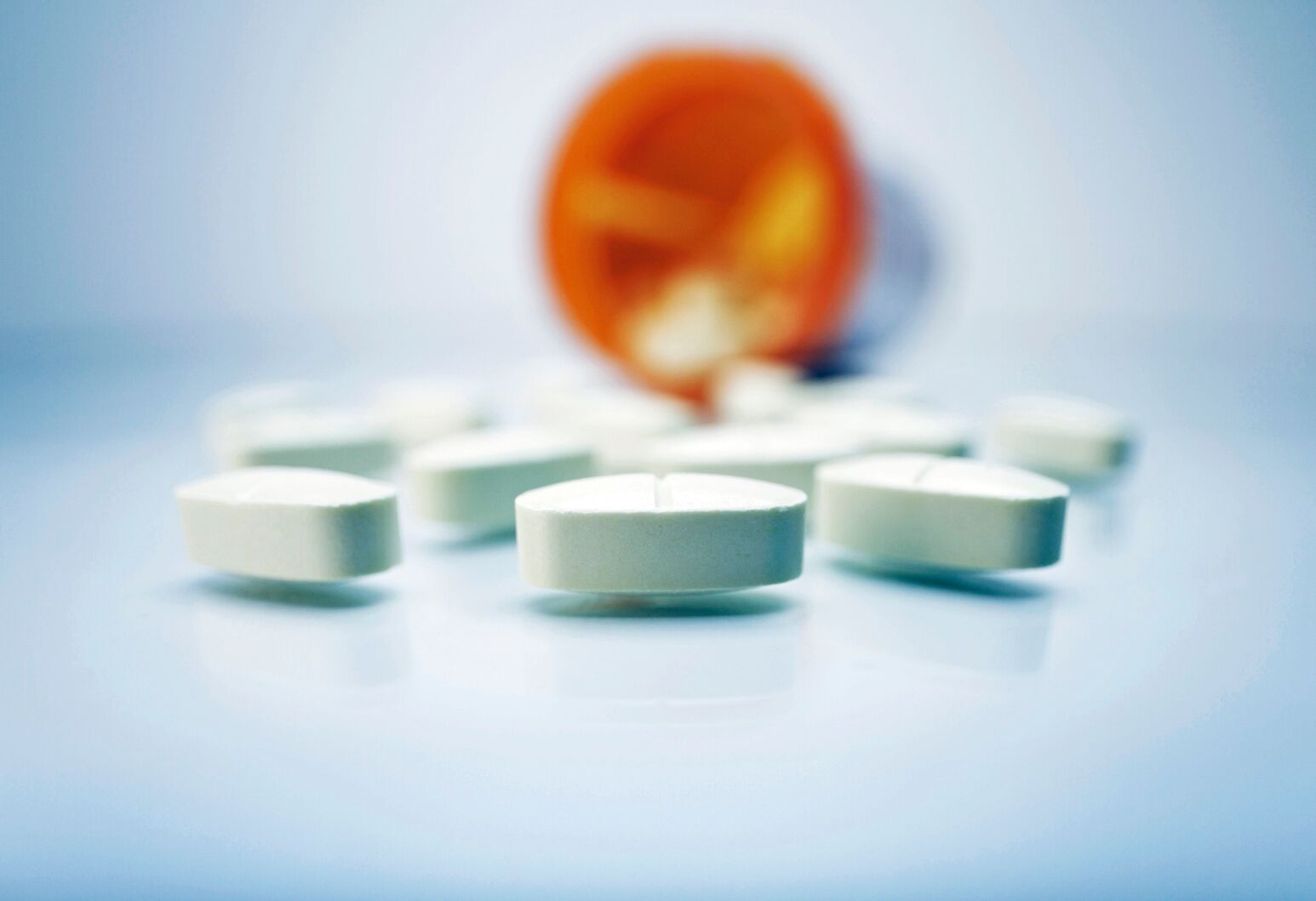 A pill bottle out of focus in the background lays on its side with white, oval tablets spilled across the bluish-white surface. 