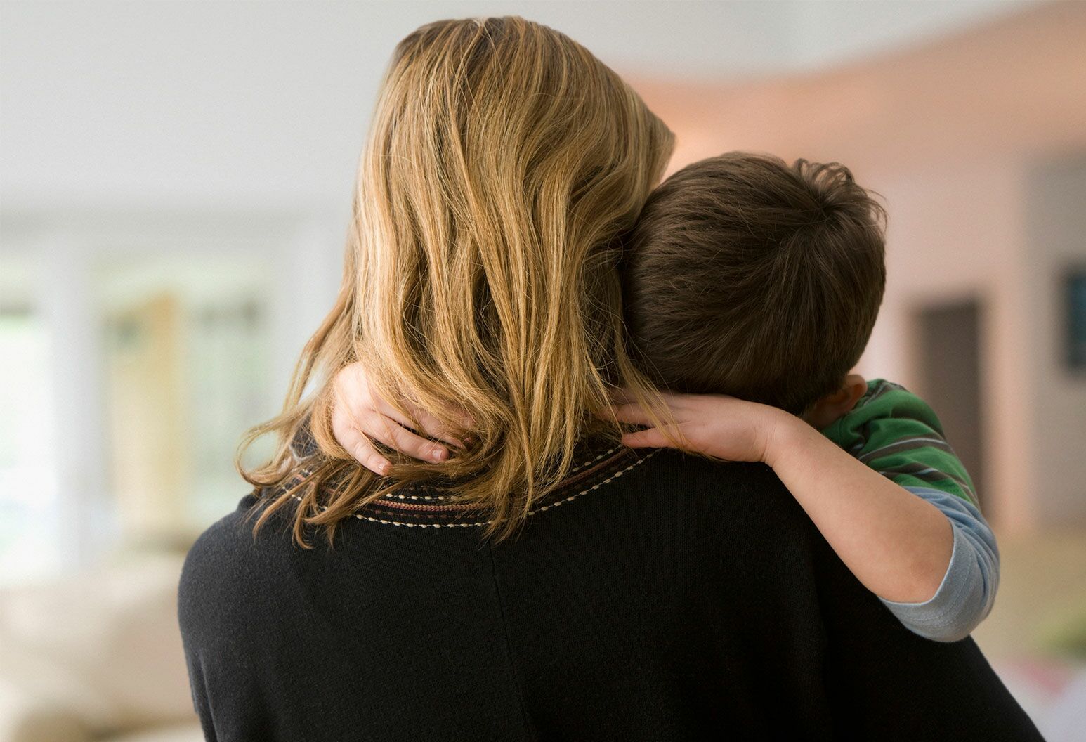 A young boy is being held by an adult woman with long blonde hair in a black sweater. His face is buried into her shoulder.