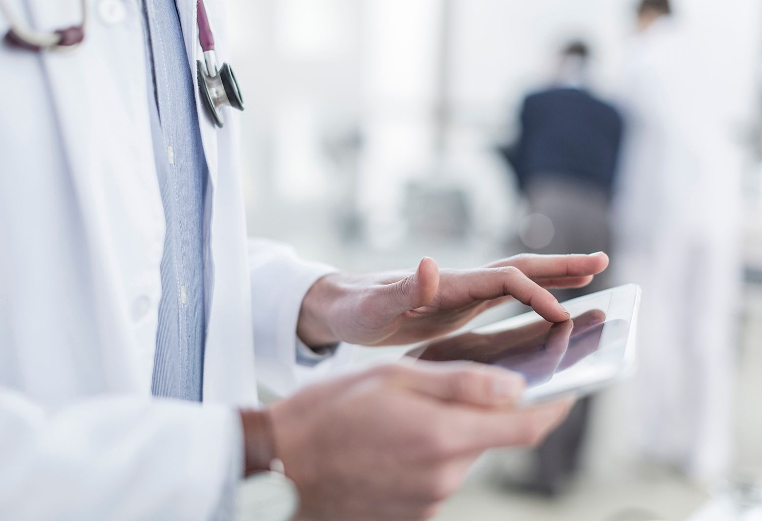 Close-up of a doctor's hands using a white tablet. He is in a white coat and wears a stethoscope.
