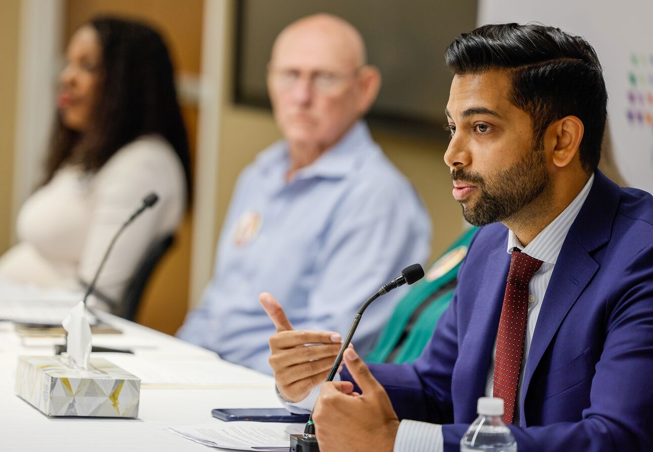 A man wearing a suit speaks into a microphones while others sitting at the same table look on.