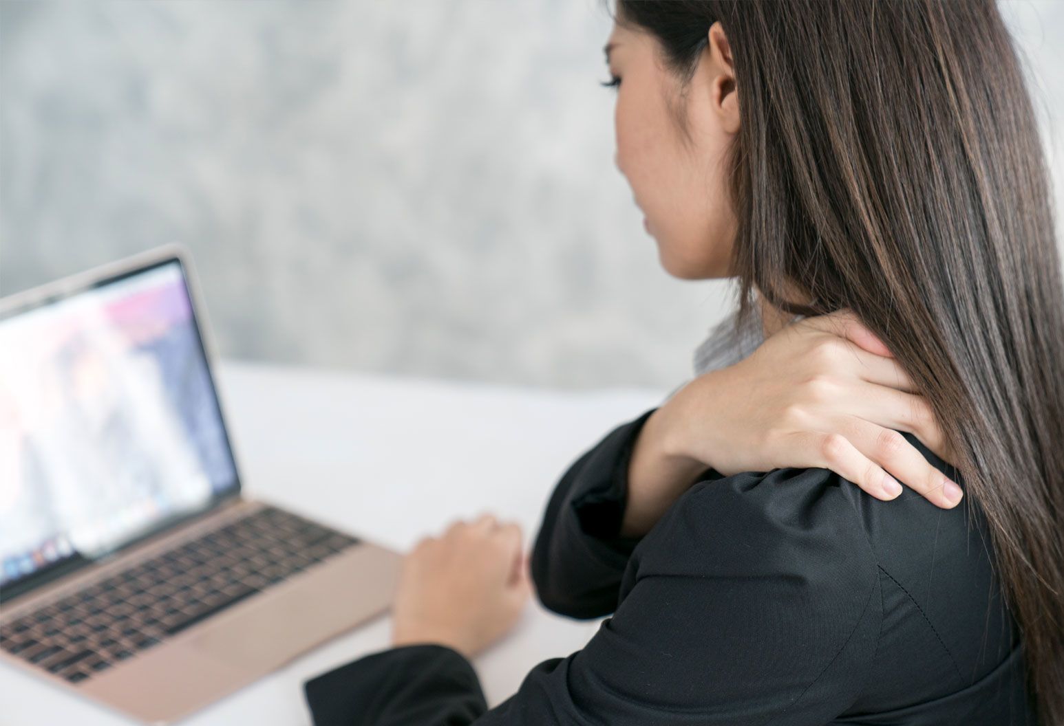 A young woman sits at a laptop and is holding her shoulder as if she is in pain. She's wearing a black shirt and has long brown hair.