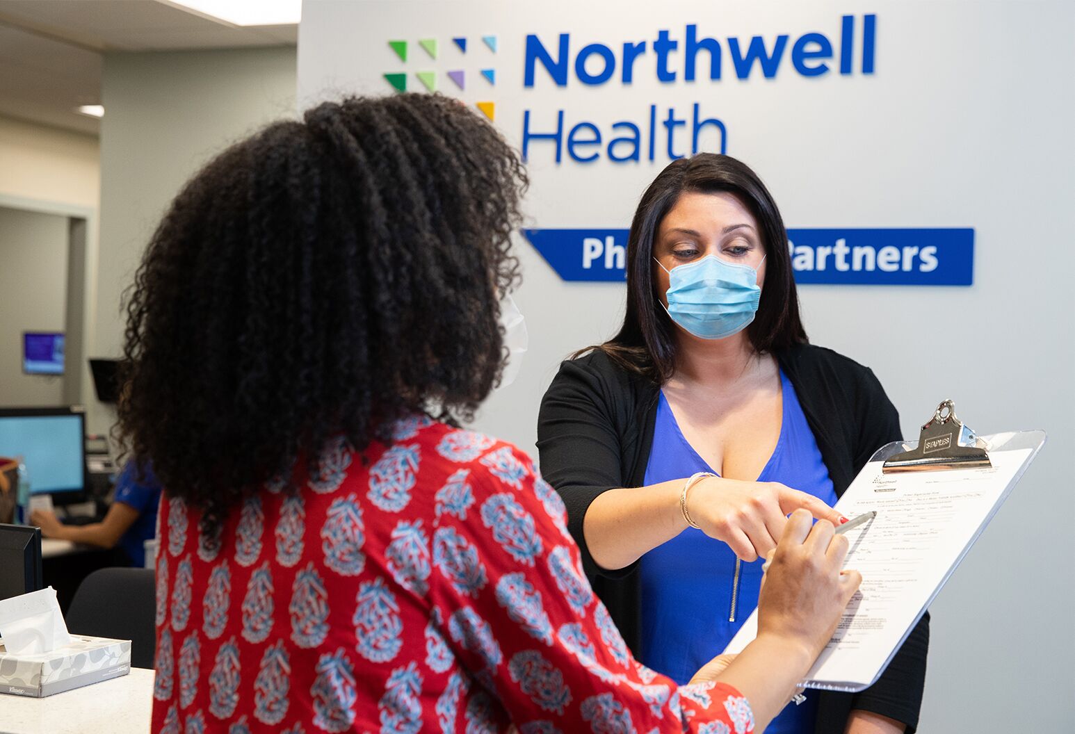 A provider in a face mask points to information on a clipboard that a patient is holding.