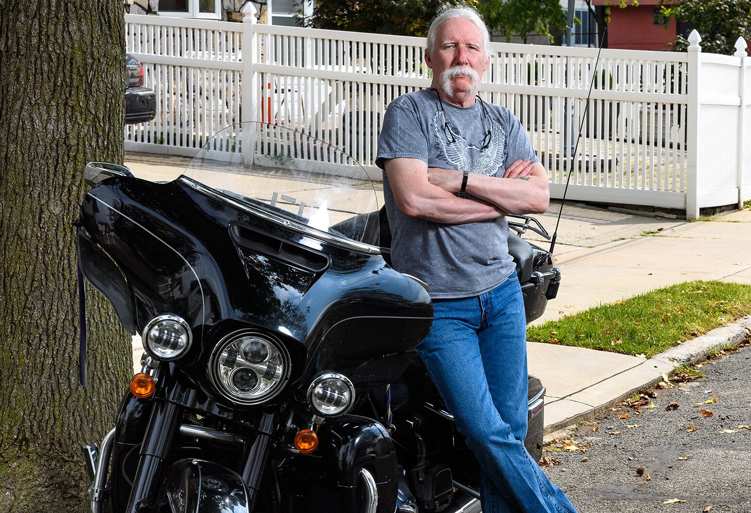Man in his 60s with a white mustache, blue jeans and a gray shirt leans on a motorcycle.
