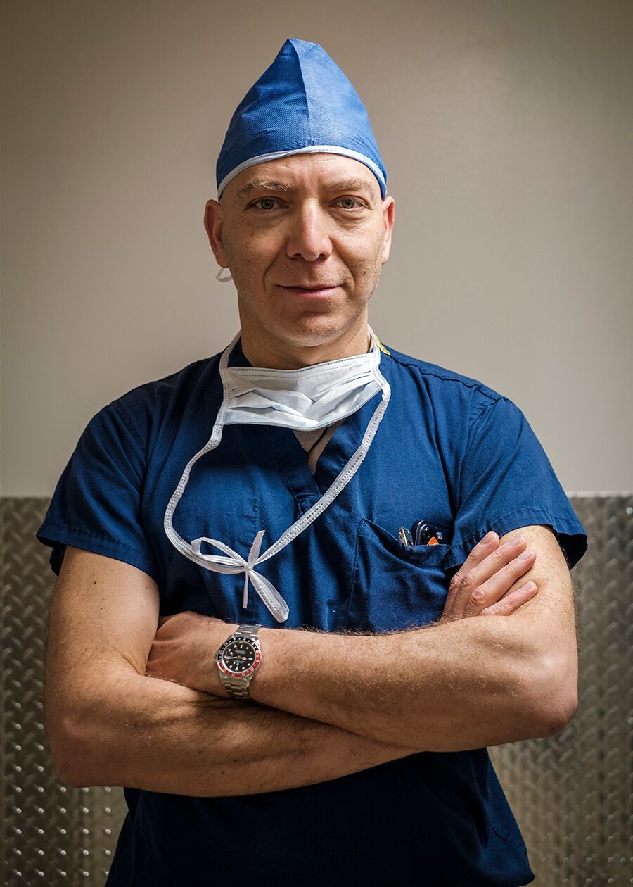 A male surgeon stands with arms folded wearing blue scrubs and head cap with a surgical face mask draped over his chest. 